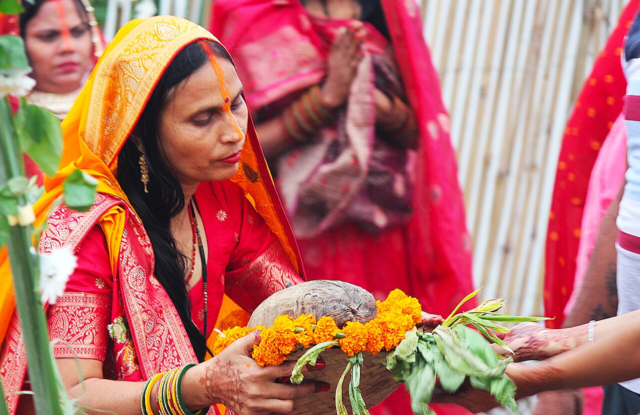 A devotee in a red and gold sari holds a bamboo soop basket with a coconut and marigolds while offering arghya during Chhath celebrations.