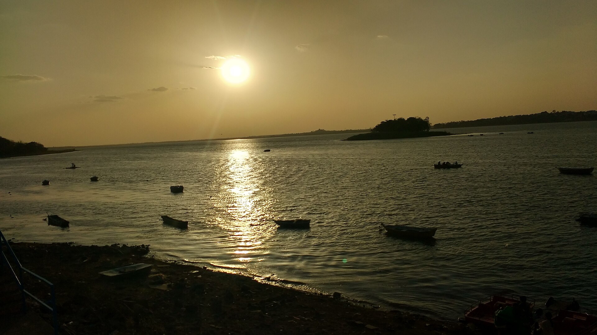 Sunset over Bhopal's Upper Lake (Bada Talaab): small boats on calm water with a wooded island and the far shoreline in silhouette.